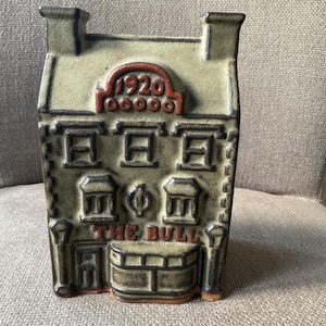 May include: A ceramic decorative piece shaped like a building, with the text "THE BULL" and "1920" in a reddish-brown color. The building has windows and a textured surface, with a light green and brown color scheme.