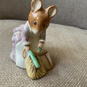 May include: A ceramic figurine of a mouse wearing a floral dress and apron, holding a green broom. The mouse has brown fur and is bent over as if sweeping. The figurine is a collectible item.