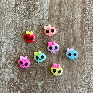 May include: Assortment of colorful skull-shaped charms with bows. The charms are made of plastic and have a glossy finish. The colors include pink, blue, green, yellow, and red.