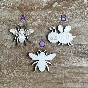 May include: Three wooden bee cutouts, one with a simple design, one with a smiling face, and one with a more detailed design. The cutouts are on a brown textured surface.
