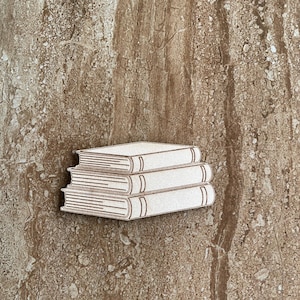 May include: A stack of three wooden books with white pages and brown outlines. The books are arranged in a neat pile, with the spines facing outward. The background is a textured brown surface.