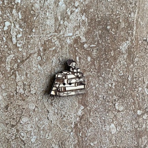May include: A wooden button featuring a person sitting atop a stack of books. The button is made of light-colored wood with dark brown etched details. The person is reading a book, and the books are stacked in a neat pile.