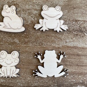 May include: Four unfinished wooden frog cutouts. The frogs are in various poses, with some facing forward and others facing sideways. The cutouts are made of light brown wood and have a smooth surface.