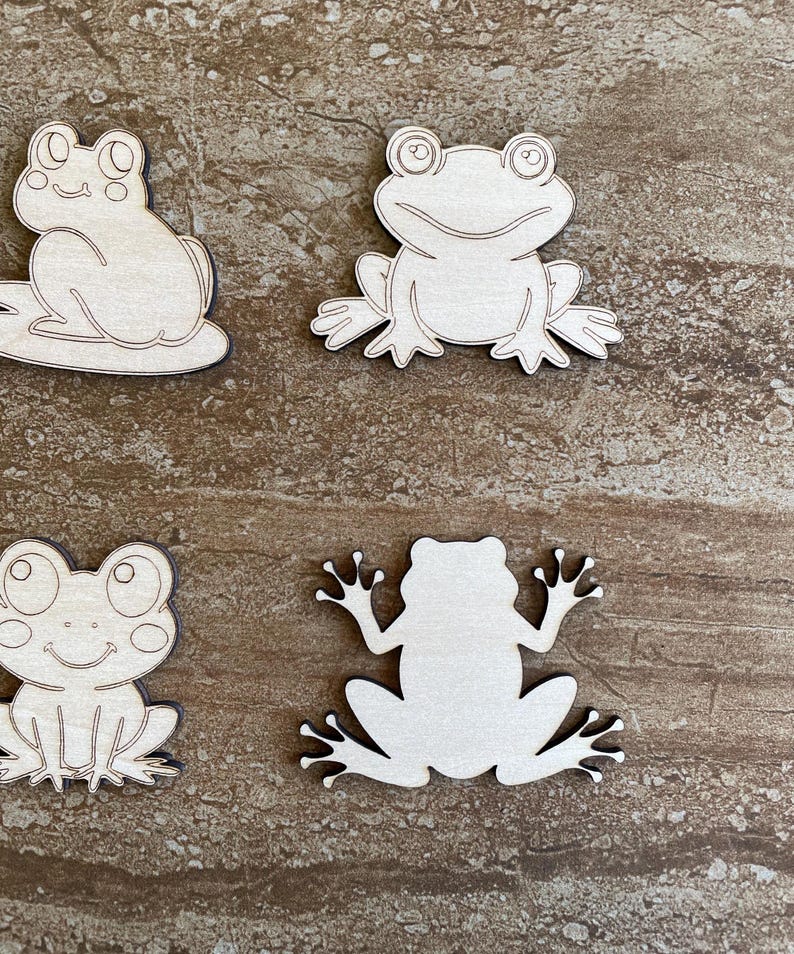 May include: Four unfinished wooden frog cutouts. The frogs are in various poses, with some facing forward and others facing sideways. The cutouts are made of light brown wood and have a smooth surface.