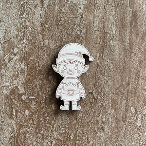 May include: A wooden cutout of a cartoon elf with a hat, pointed ears, and a belt. The elf is light brown with etched details, standing upright. The background is a textured, mottled brown.