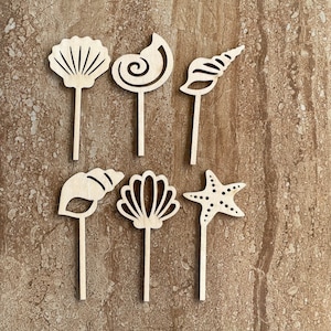 May include: Seven wooden beach-themed decorative picks. The set includes a starfish, a scallop shell, a spiral shell, and other seashell designs. Each pick has a long, thin stem. The items are made of light-colored wood.