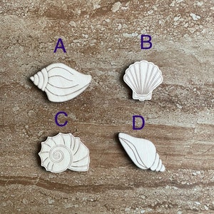 May include: Four different wooden cutout shapes of seashells. The shells are labeled A, B, C, and D.  The shells are all white and have a natural wood grain.