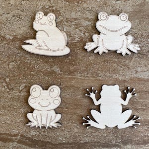 May include: Four unfinished wooden frog cutouts. Two frogs are sitting with their legs tucked in, and two frogs are sitting with their legs spread out. The frogs are all facing forward.