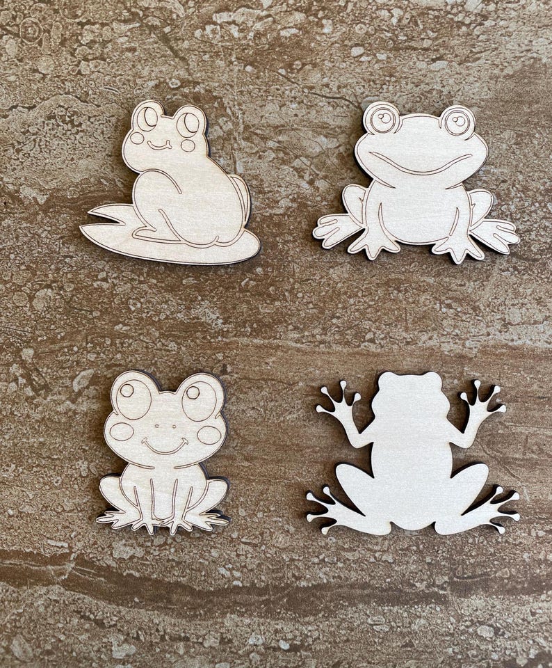 May include: Four unfinished wooden frog cutouts. Two frogs are sitting with their legs tucked in, and two frogs are sitting with their legs spread out. The frogs are all facing forward.