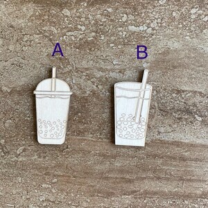 May include: Two wooden cutout designs of bubble tea with straws. The design on the left has a lid and the design on the right is a simple cup. Both designs are white against a brown background.