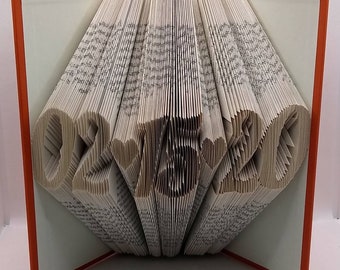 May include: A book with orange covers featuring folded pages that form the date 02-15-20 with heart shapes between the numbers. The pages are precisely creased to create the three-dimensional numeric and heart designs visible from the front.