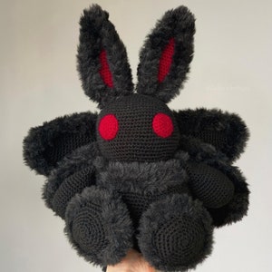 Op de afbeelding: Een zwart gehaakt amigurumi-speelgoed met rode ogen en lange, slappe oren. Het speelgoed wordt in de hand van een persoon gehouden. Het speelgoed is een digitaal haakpatroon.