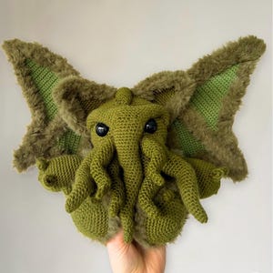 以下が含まれることがあります： 大きな目と触手を持つ緑色の編みぐるみクトゥルフ。ぬいぐるみは手で持たれています。