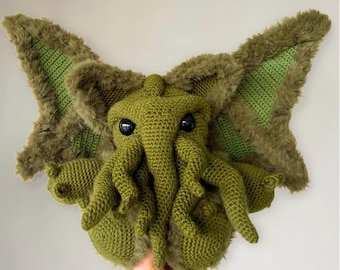 May include: Green crocheted Cthulhu plush toy with large eyes and tentacles. The plush toy is being held by a hand.