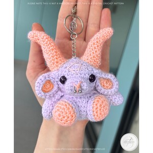 May include: A crocheted lavender goat keychain with peach-colored horns, ears, and feet. The goat has black eyes, a silver nose ring, and a pentagram on its head. The keychain is attached to a silver ring. The text in the image says "PLEASE NOTE THIS IS NOT A PHYSICAL PRODUCT, THIS IS A DIGITAL CROCHET PATTERN."