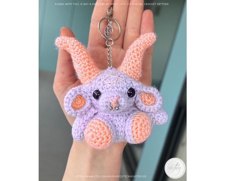 May include: A crocheted lavender goat keychain with peach-colored horns, ears, and feet. The goat has black eyes, a silver nose ring, and a pentagram on its head. The keychain is attached to a silver ring. The text in the image says "PLEASE NOTE THIS IS NOT A PHYSICAL PRODUCT, THIS IS A DIGITAL CROCHET PATTERN."