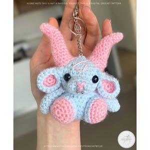 May include: A crocheted keychain featuring a light blue and pink goat-like creature with black eyes and a silver nose ring. The keychain has a silver chain and ring. The text "PLEASE NOTE THIS IS NOT A PHYSICAL PRODUCT, THIS IS A DIGITAL CROCHET PATTERN" is visible.