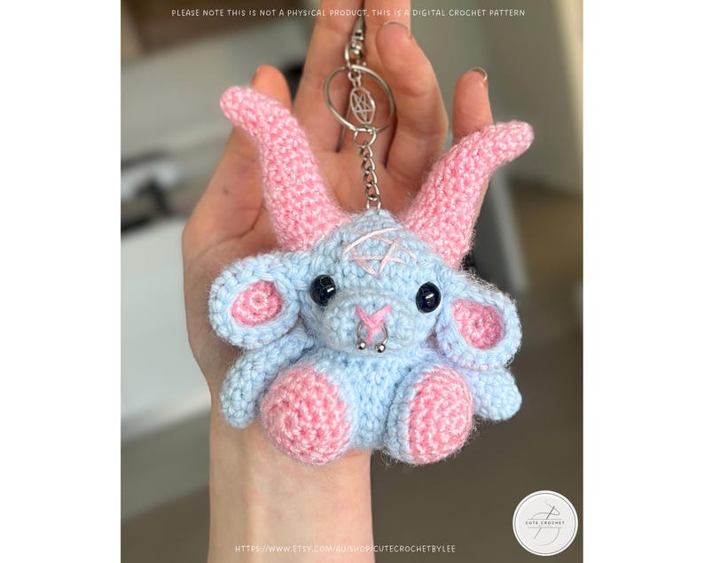 May include: A crocheted keychain featuring a light blue and pink goat-like creature with black eyes and a silver nose ring. The keychain has a silver chain and ring. The text "PLEASE NOTE THIS IS NOT A PHYSICAL PRODUCT, THIS IS A DIGITAL CROCHET PATTERN" is visible.