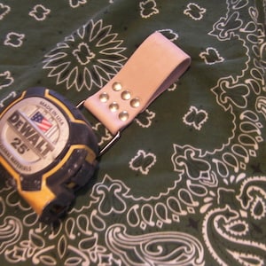 May include: A yellow and black DeWalt 25' tape measure with a pink leather belt loop with metal rivets. The tape measure is on a green and white bandana.