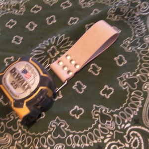 May include: A yellow and black tape measure with the text "DEWALT 25'" on the face, attached to a light pink leather strap with silver metal rivets.