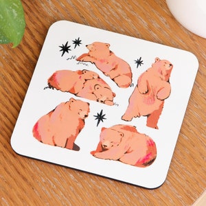 Könnte beinhalten: Quadratischer weißer Untersetzer mit Illustrationen von braunen Bären in verschiedenen Posen und schwarzen Sternakzenten. Der Untersetzer liegt auf einem Holztisch, in der Nähe einer weißen Tasse und einer grünen Pflanze.