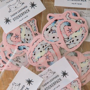 Puede incluir: Una colección de pegatinas de leopardo de las nieves rosas y etiquetas de paquetes de pegatinas blancas. Las pegatinas presentan una ilustración de leopardo de las nieves con manchas azules y negras. Las etiquetas dicen "SNOW LEOPARD STICKERPACK" con un diseño de estrella.