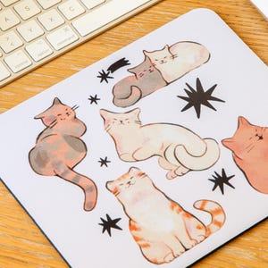 May include: White mousepad featuring watercolor illustrations of various cats in different poses, with black star accents. A computer keyboard and mouse are in the background, suggesting a desk setup. The cats are in shades of orange, gray, and white.