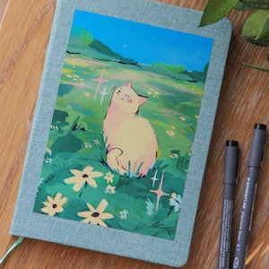 Puede incluir: Un diario azul claro con una ilustraci&oacute;n de un gato en la portada. La ilustraci&oacute;n representa un gato en un campo de flores. Dos bol&iacute;grafos negros est&aacute;n colocados sobre la superficie de madera junto al diario.