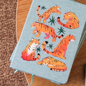 May include: A stack of light blue notebooks with orange tiger illustrations and green star accents. The tigers are in various poses. A light blue ribbon is attached to the top notebook.