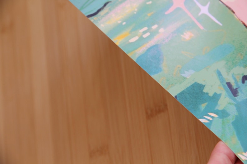 May include: A section of a colorful abstract art print with a light brown wooden surface in the background. The print features a mix of teal, blue, pink, and yellow brushstrokes, with star-like shapes.
