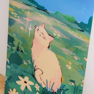 May include: An art print featuring a cream-colored cat basking in sunlight on a grassy hill. The illustration has a whimsical style with a blue sky, green grass, and white flowers. The artist's signature "MADI" is in the lower left corner.