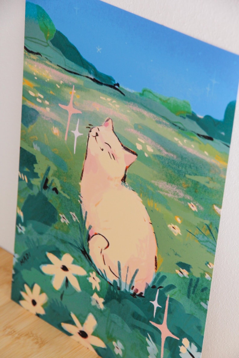 May include: An art print featuring a cream-colored cat basking in sunlight on a grassy hill. The illustration has a whimsical style with a blue sky, green grass, and white flowers. The artist's signature "MADI" is in the lower left corner.