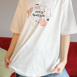 Puede incluir: Camiseta color crema con una ilustraci&oacute;n caprichosa de gatos y el texto "stay Pawsitive". El dise&ntilde;o incluye varios gatos de dibujos animados en tonos marr&oacute;n, naranja y rosa. La camiseta tiene mangas cortas y un corte holgado.