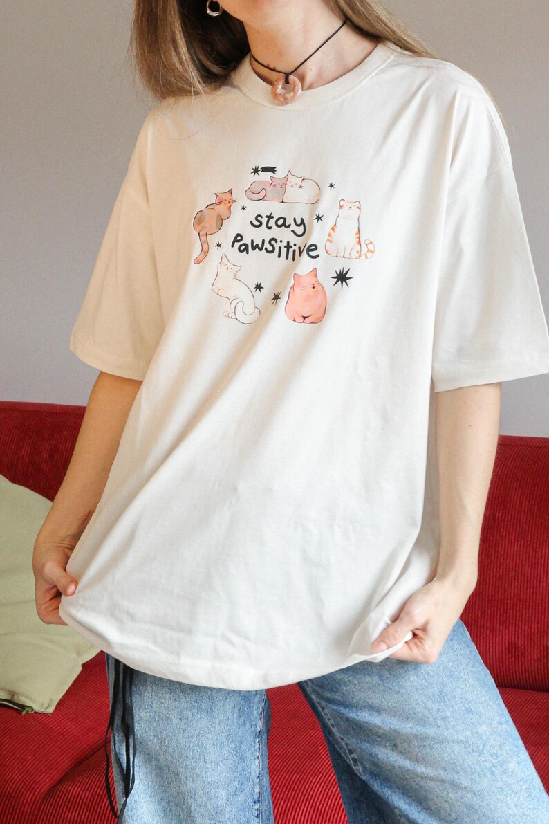 Puede incluir: Camiseta color crema con una ilustraci&oacute;n caprichosa de gatos y el texto "stay Pawsitive". El dise&ntilde;o incluye varios gatos de dibujos animados en tonos marr&oacute;n, naranja y rosa. La camiseta tiene mangas cortas y un corte holgado.