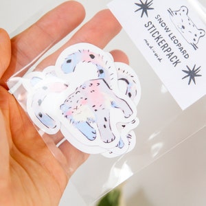 Puede incluir: Una bolsa de pl&aacute;stico transparente que contiene un paquete de pegatinas con ilustraciones de leopardos de las nieves. Las pegatinas son blancas con dise&ntilde;os de acuarela rosa y azul. La bolsa tiene una etiqueta que dice "Snow Leopard Stickerpack" con un dise&ntilde;o de estrella.