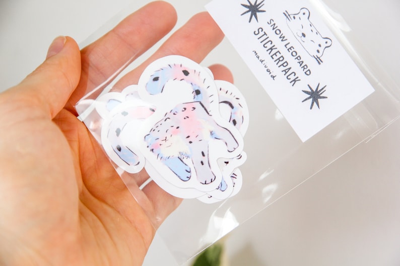 Puede incluir: Una bolsa de pl&aacute;stico transparente que contiene un paquete de pegatinas con ilustraciones de leopardos de las nieves. Las pegatinas son blancas con dise&ntilde;os de acuarela rosa y azul. La bolsa tiene una etiqueta que dice "Snow Leopard Stickerpack" con un dise&ntilde;o de estrella.