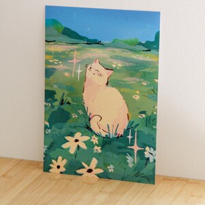 May include: A whimsical art print featuring a light-colored cat sitting in a field of flowers under a starry night sky. The illustration has a soft color palette with green, blue, and yellow hues. The artist's signature "MADI" is visible.