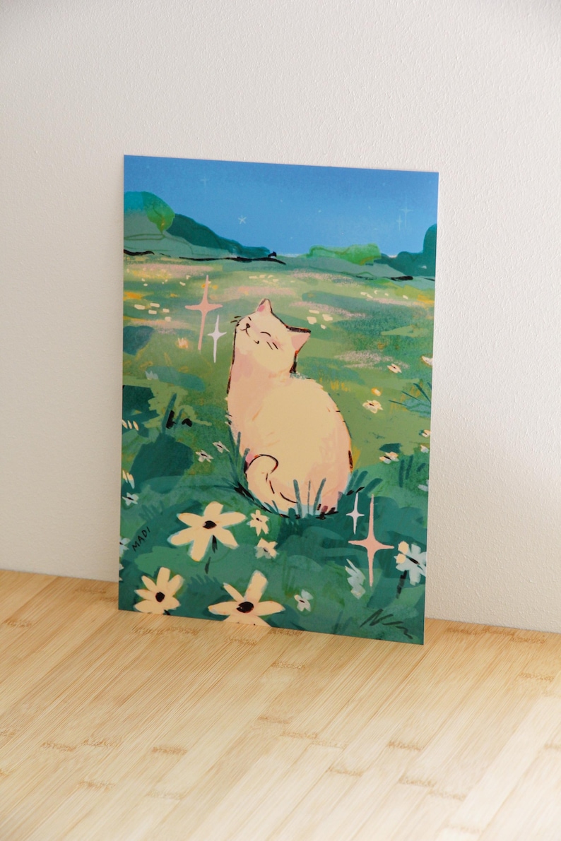 May include: A whimsical art print featuring a light-colored cat sitting in a field of flowers under a starry night sky. The illustration has a soft color palette with green, blue, and yellow hues. The artist's signature "MADI" is visible.