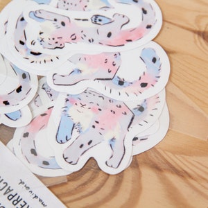 Puede incluir: Una hoja de pegatinas con ilustraciones de acuarela de un gato manchado en varias poses. El gato est&aacute; representado en tonos de rosa, azul y gris.