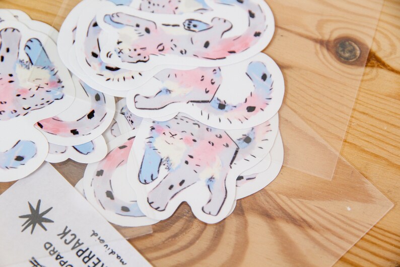 Puede incluir: Una hoja de pegatinas con ilustraciones de acuarela de un gato manchado en varias poses. El gato est&aacute; representado en tonos de rosa, azul y gris.