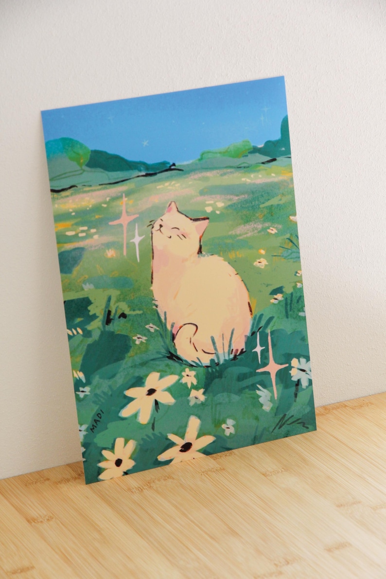 May include: A print featuring a smiling cat in a field of flowers under a blue sky. The cat is light-colored, and the flowers are white and yellow. The artwork has a whimsical, illustrative style.