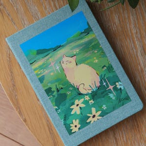 Puede incluir: Un diario azul claro con una colorida ilustraci&oacute;n de un gato en un campo de flores. El gato es blanco y las flores son amarillas y blancas. El diario est&aacute; sobre una superficie de madera.