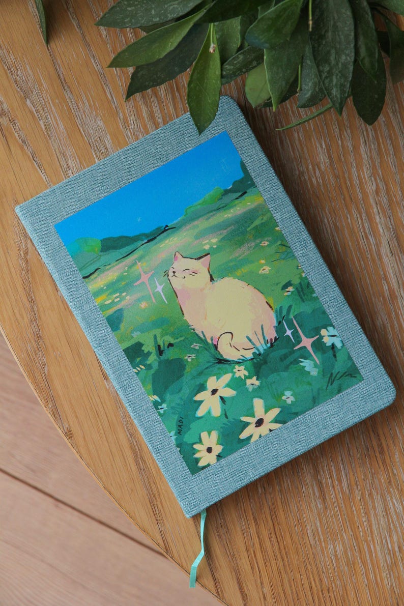 Puede incluir: Un diario azul claro con una colorida ilustraci&oacute;n de un gato en un campo de flores. El gato es blanco y las flores son amarillas y blancas. El diario est&aacute; sobre una superficie de madera.
