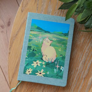 Puede incluir: Un diario azul claro con una colorida ilustraci&oacute;n de un gato en un campo de flores. El gato es blanco con acentos rosados, y el fondo presenta un cielo azul y hierba verde. El diario est&aacute; sobre una mesa de madera.
