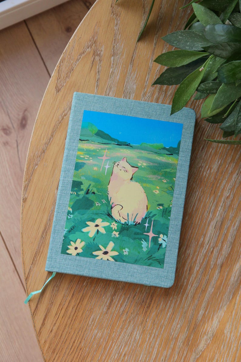 Puede incluir: Un diario azul claro con una colorida ilustraci&oacute;n de un gato en un campo de flores. El gato es blanco con acentos rosados, y el fondo presenta un cielo azul y hierba verde. El diario est&aacute; sobre una mesa de madera.