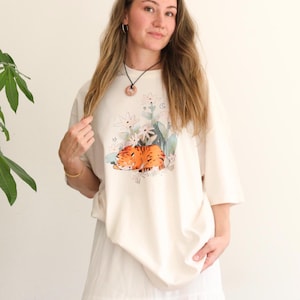 May include: Cream-colored t-shirt featuring a graphic of a sleeping orange tiger surrounded by flowers and foliage. The shirt has short sleeves and a relaxed fit. The person is wearing a white skirt and a necklace.