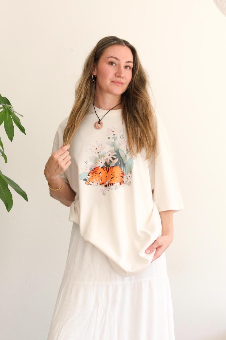 May include: Cream-colored t-shirt featuring a graphic of a sleeping orange tiger surrounded by flowers and foliage. The shirt has short sleeves and a relaxed fit. The person is wearing a white skirt and a necklace.