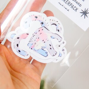 Puede incluir: Un conjunto de cuatro pegatinas con una ilustraci&oacute;n acuarela rosa y azul de un leopardo. Las pegatinas est&aacute;n empaquetadas en una bolsa de pl&aacute;stico transparente con una etiqueta blanca que dice "Stickerpack madivoid".