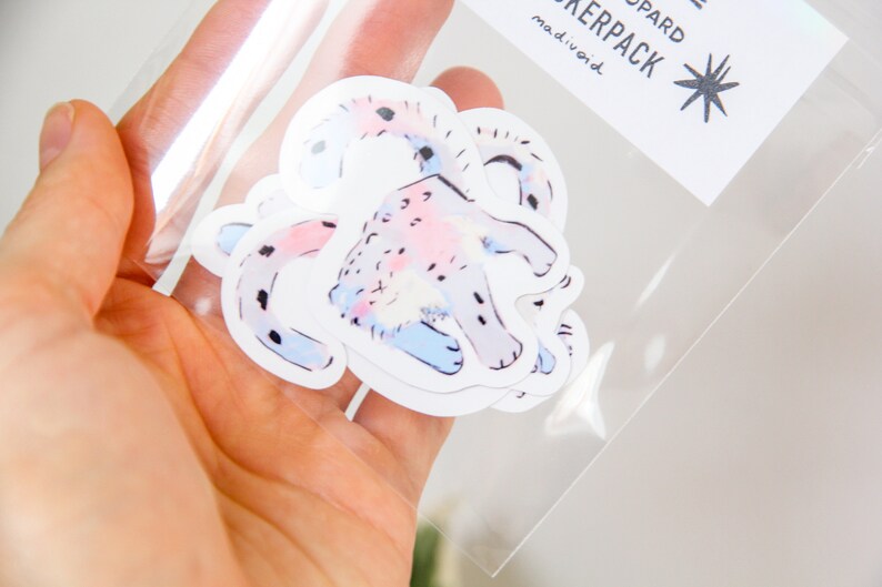 Puede incluir: Un conjunto de cuatro pegatinas con una ilustraci&oacute;n acuarela rosa y azul de un leopardo. Las pegatinas est&aacute;n empaquetadas en una bolsa de pl&aacute;stico transparente con una etiqueta blanca que dice "Stickerpack madivoid".