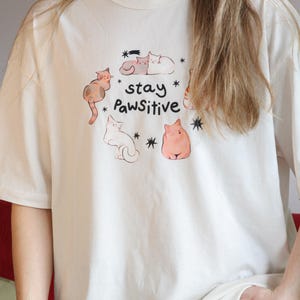Puede incluir: Camiseta de color crema con un dise&ntilde;o l&uacute;dico de gatos de dibujos animados en varias poses. La camiseta incluye el texto "stay Pawsitive" con acentos de estrellas. Una parte superior informal y c&oacute;moda.
